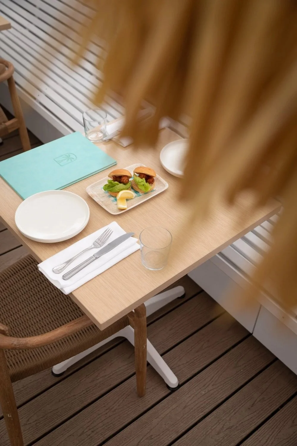 A table set with a white plate, a fork, a knife, a glass, a napkin, and a plate with small sandwiches, lemon slice, and greens. There is a wooden chair and a blue menu or notebook on the table.