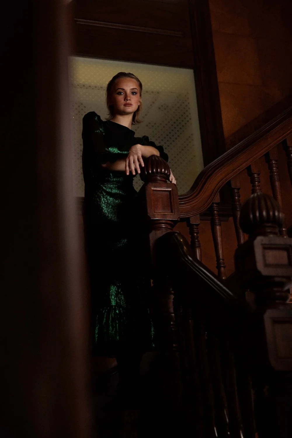 Woman in a black and green dress standing on a staircase, leaning on the wooden handrail, in a dimly lit interior.
