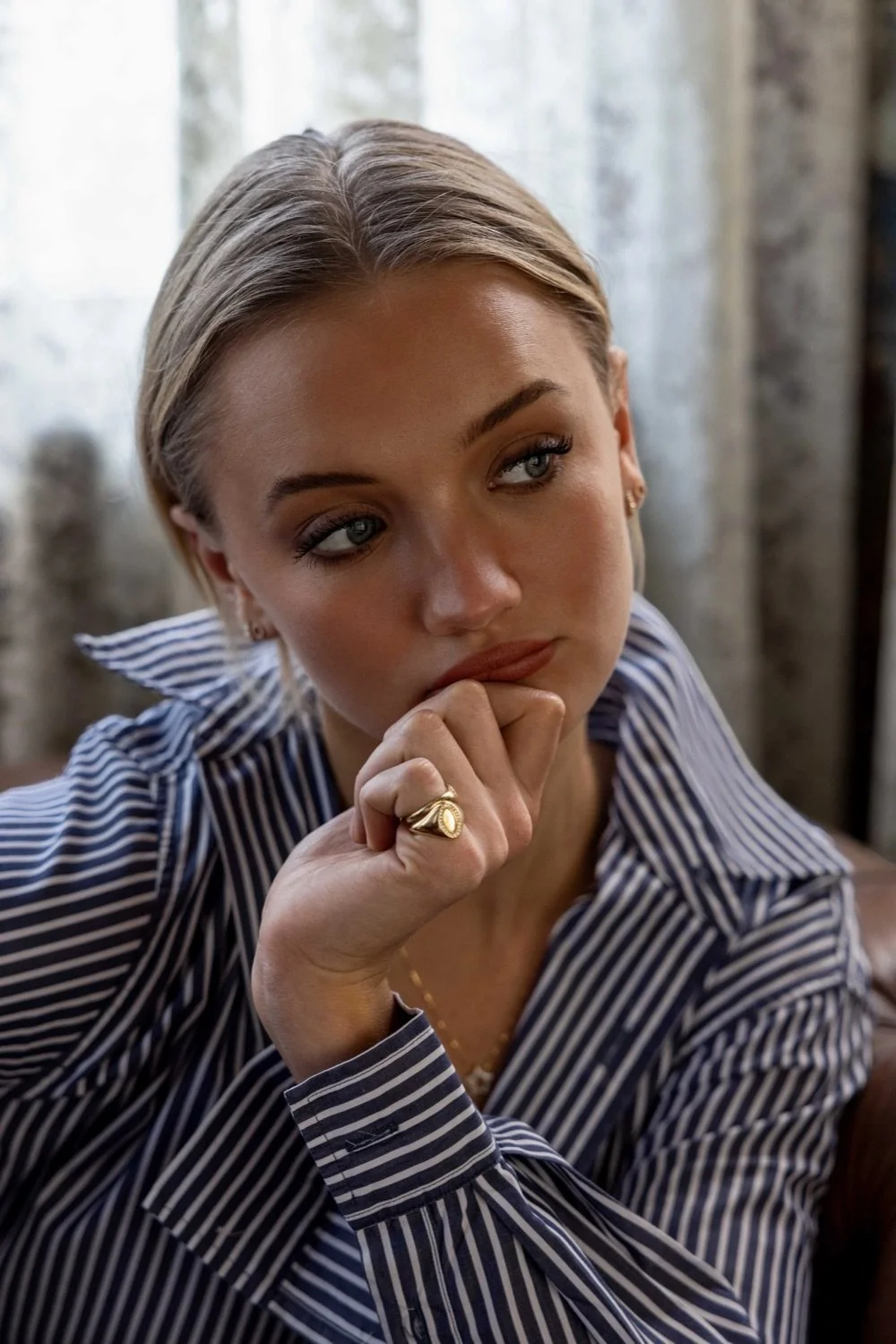 A woman with blonde hair, styled straight, wearing a striped shirt, sitting indoors near a window with a thoughtful expression, resting her chin on her hand and looking to the side.