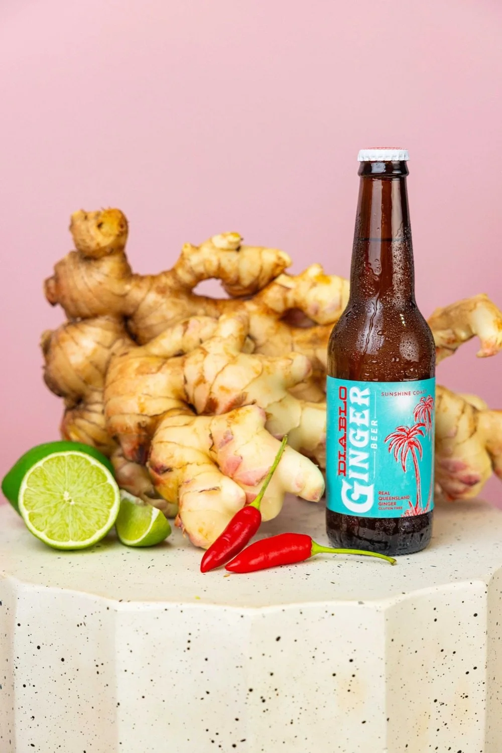 A bottle of ginger beer with a turquoise label, fresh ginger roots, a lime cut in half, and two small red chili peppers arranged on a speckled white surface against a pink background.