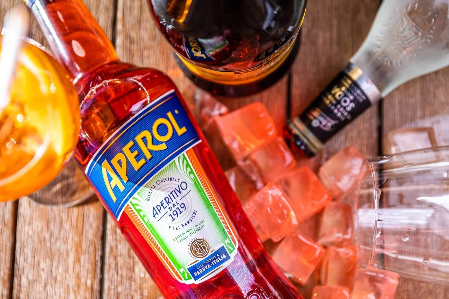 A bottle of Aperol with ice cubes and other bottles on a wooden surface.