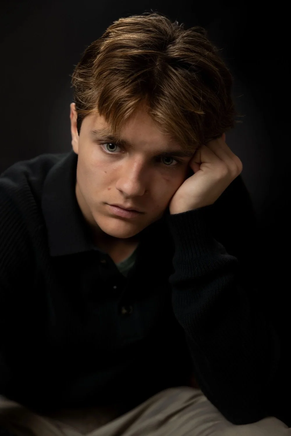 Portrait of a young man with brown hair and blue eyes resting his head on his hand, against a dark background.