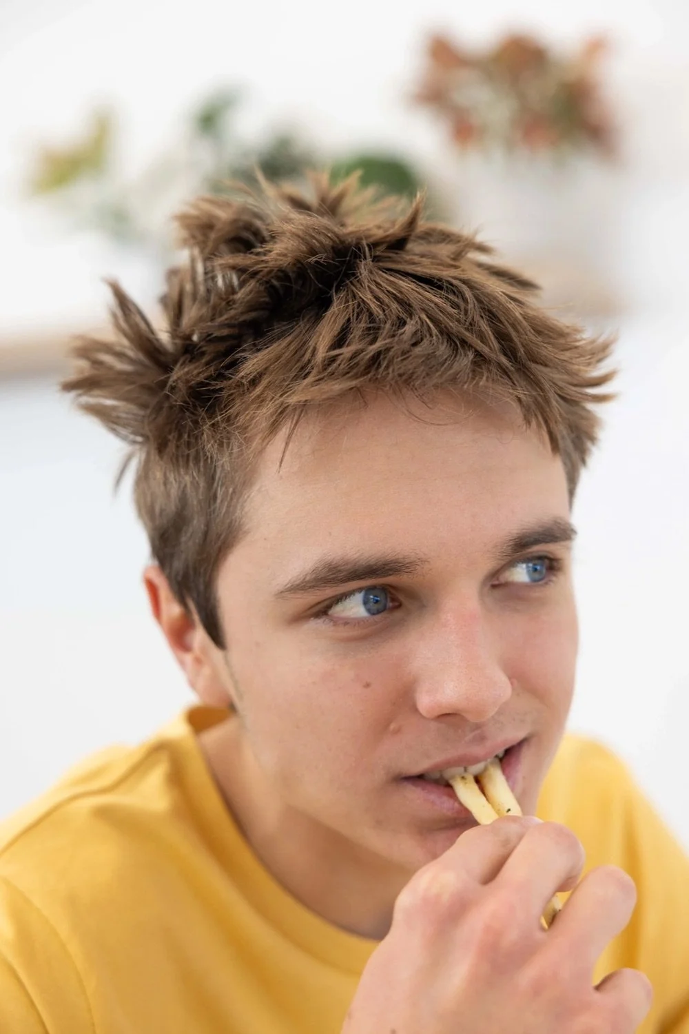 A young man with spiky, tousled brown hair and blue eyes eating French fries, wearing a yellow shirt.