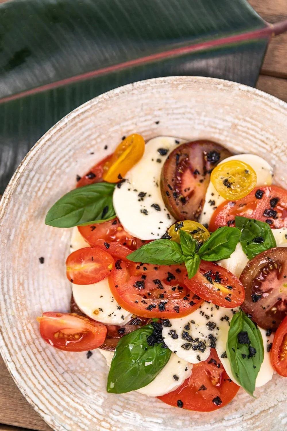 A Caprese salad with sliced heirloom tomatoes, fresh mozzarella, basil leaves, black pepper, and balsamic vinegar on a ceramic plate.