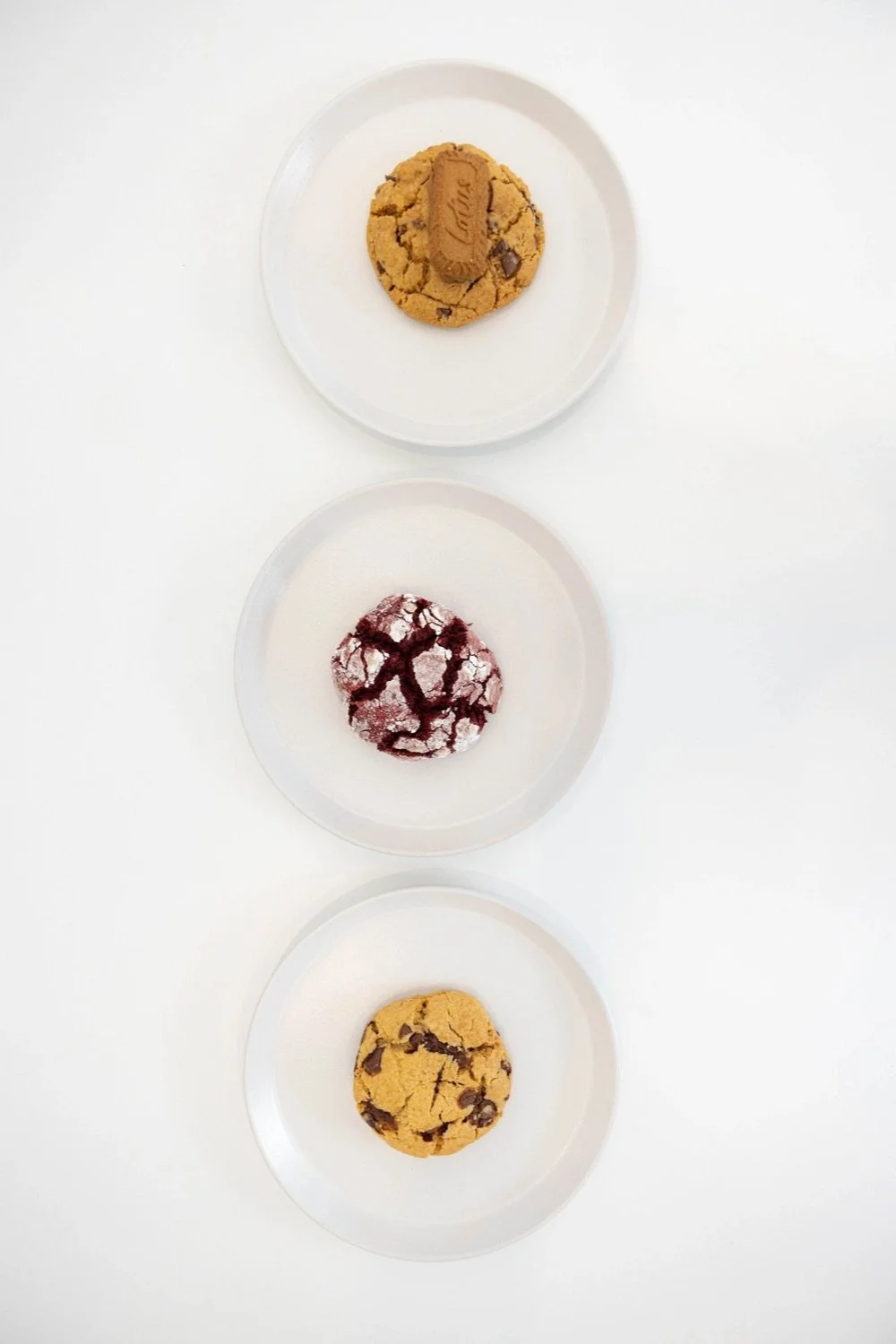 Three plates with cookies, from top to bottom: a chocolate chip cookie with a Kinder Bueno chocolate and a piece of chocolate, a red velvet cookie with white frosting and red syrup, and a chocolate chip cookie.