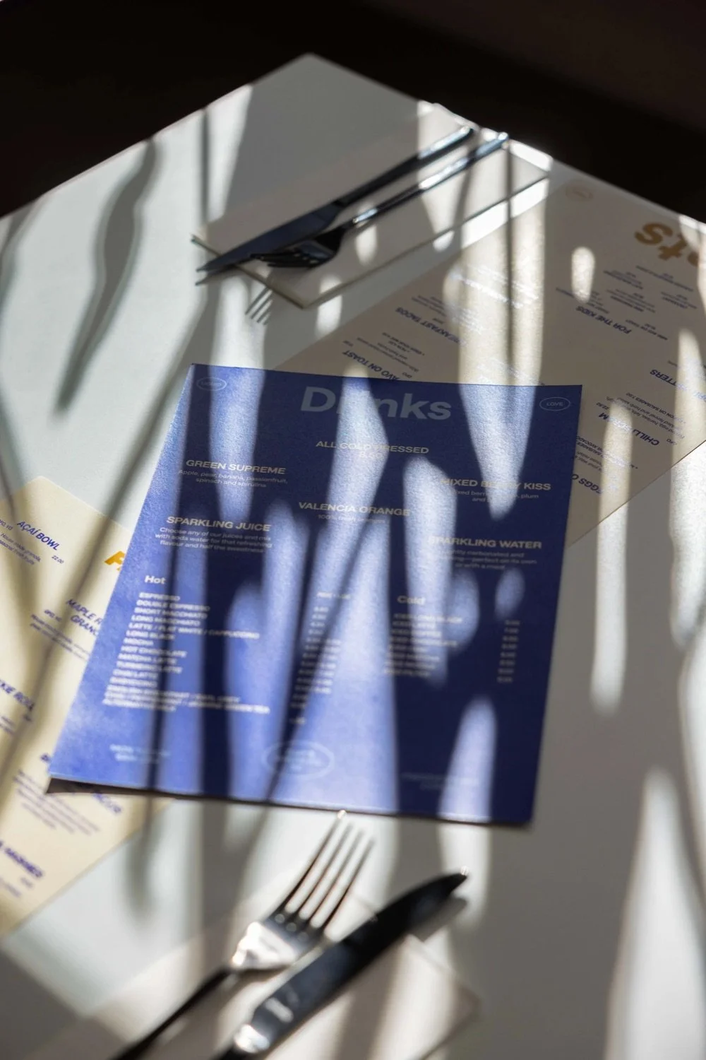 A restaurant table setting with a fork and knife, a white napkin, and menus, with sunlight and shadows cast across the table.