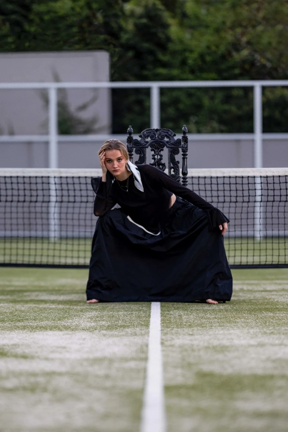 A woman dressed in black, sitting on a tennis court with a vintage black chair behind her, leaning forward with her hand on her face and looking into the camera.