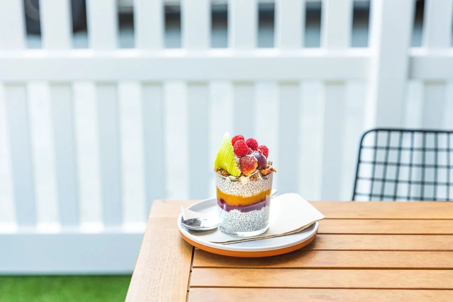 A glass with chia seed pudding topped with assorted fresh berries and green fruit slices on a plate with a spoon, set on a wooden table with a white railing backdrop.