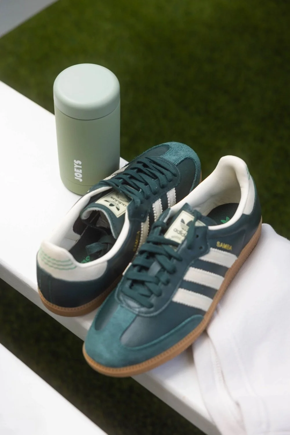 A pair of vintage-style Adidas sneakers on white pants, with a light gray water bottle labeled "JOEYS" behind them, placed on a white surface with grass in the background.