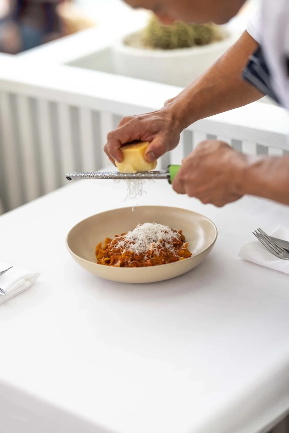 Person grating cheese over a bowl of pasta on a white table.