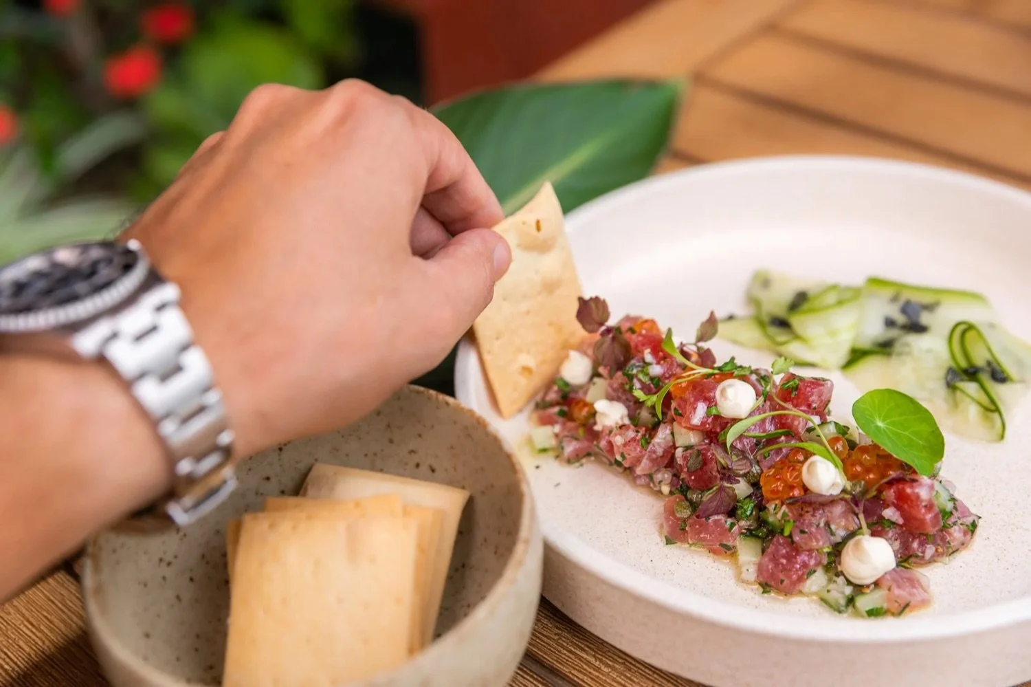 A hand is placing a crispy chip on a plate of tuna tartare with cucumber slices and garnishes, with additional chips in a bowl nearby.