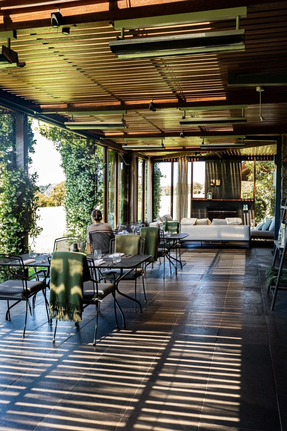 Interior of a modern restaurant with large windows, wooden ceiling, and natural sunlight, featuring tables set for dining and a woman sitting alone.