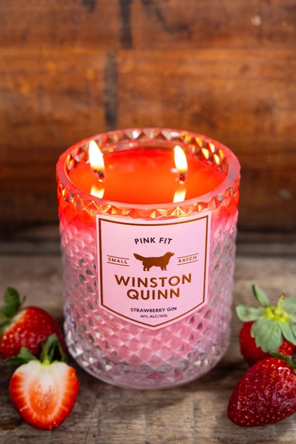 Pink glass candle holder with lit candles inside, label reads 'Pink Fit Small Batch Winston Quinn Strawberry Gin 40% Alc/Vol,' surrounded by strawberries on a wooden surface.