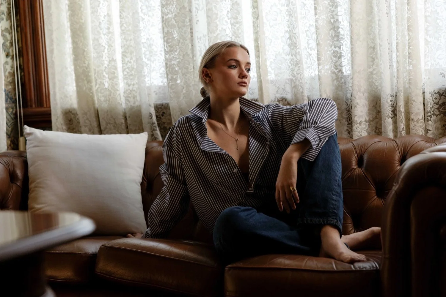 A young woman with blonde hair sitting on a brown leather couch, looking to the side with a thoughtful expression. She is wearing a striped shirt and jeans, with sheer curtains behind her.