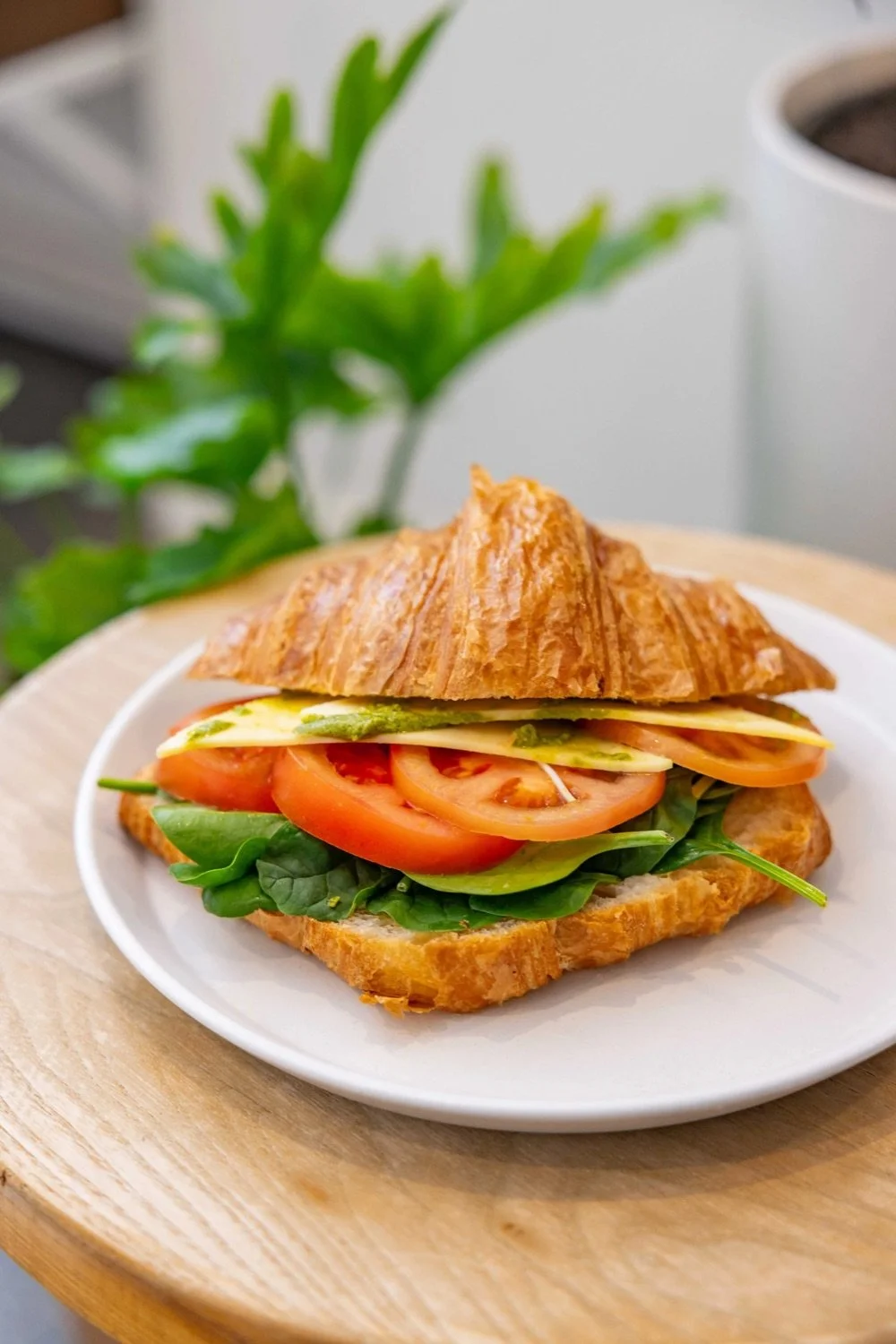 Croissant sandwich with tomato, lettuce, cheese, and avocado on a white plate.