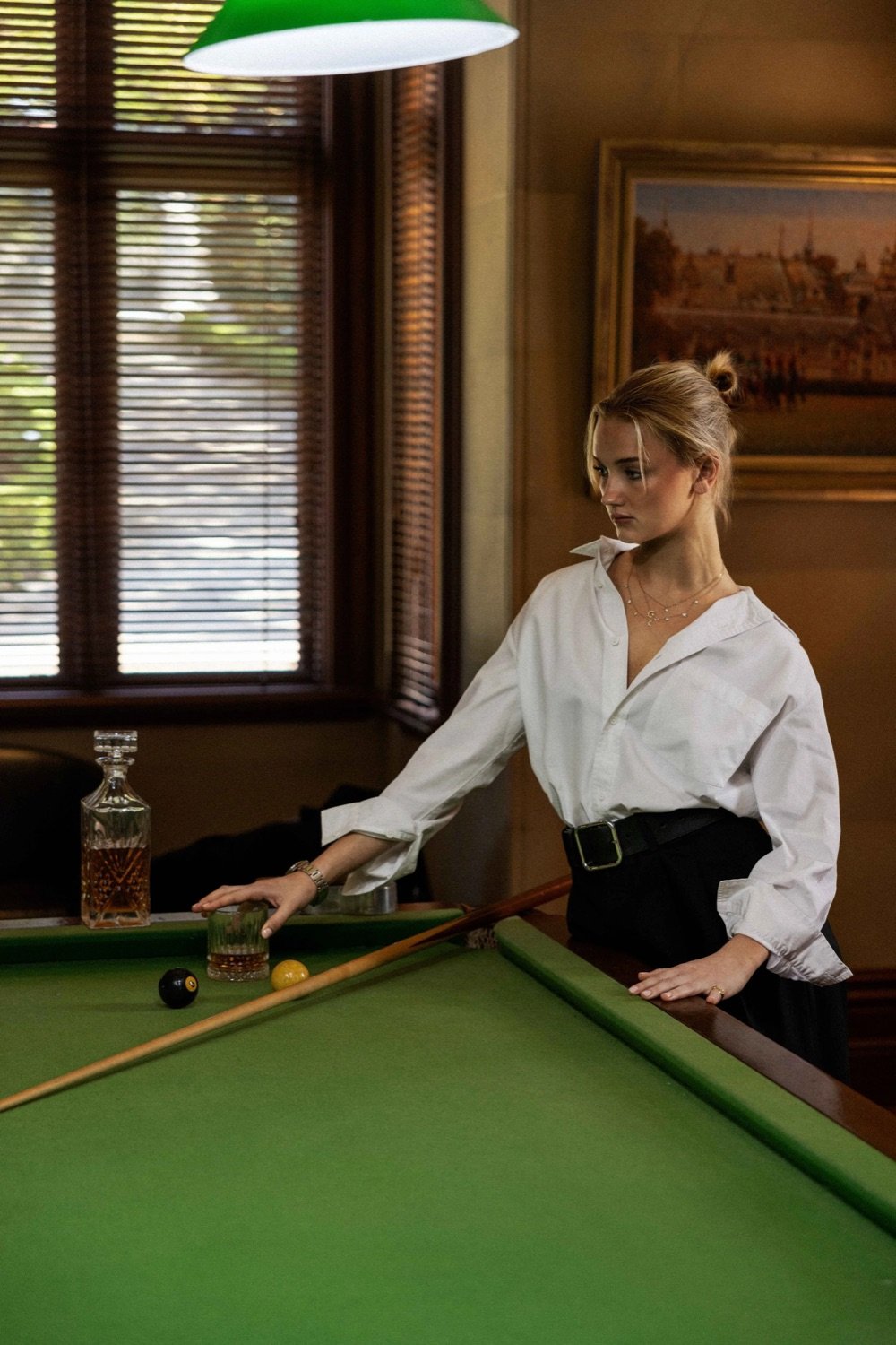 A woman with blonde hair tied back, wearing a white shirt and black pants, leaning on a green pool table with a cue stick, a glass of whiskey, a decanter, and billiard balls on the table, in a room with wooden window blinds and a landscape painting o