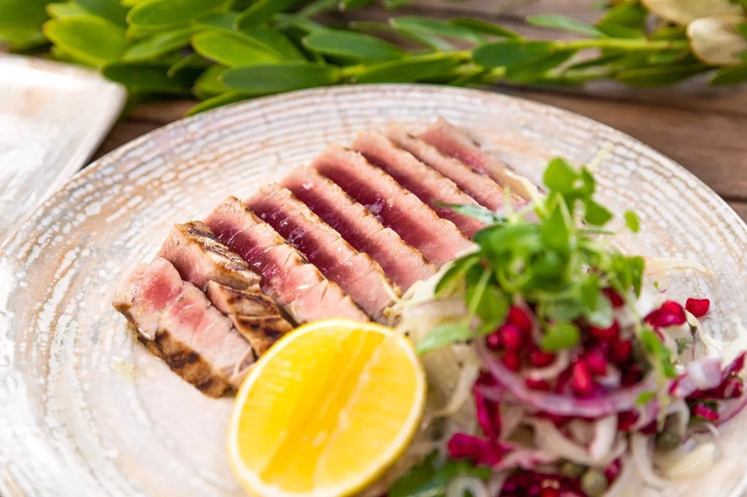 Sliced seared tuna steak with lemon wedge and salad garnished with microgreens on a ceramic plate.