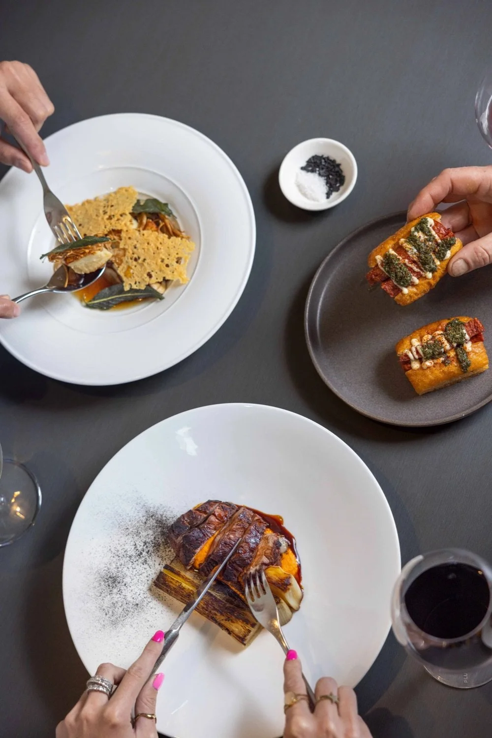 Three plates of gourmet food on a gray table, including a roasted meat with sauce, a plate with breaded appetizers topped with sauces and herbs, and a dish with a layered savory pastry. Hands with jewelry are seen holding forks and knives to cut the 