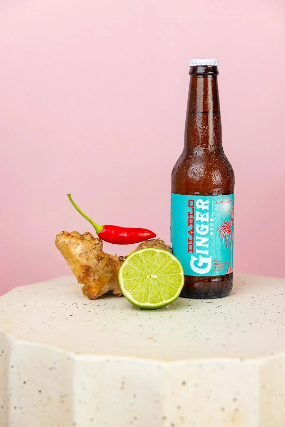 A bottle of ginger beer, a halved lime, a piece of ginger root, and a red chili pepper placed on a white textured surface against a pink background.