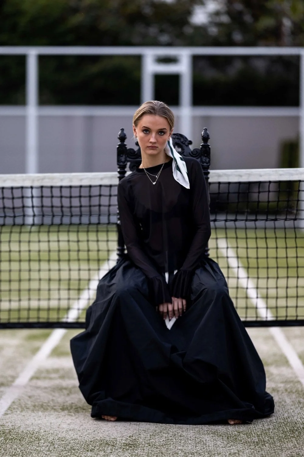 A young woman with blonde hair in a light scarf, wearing a black sheer blouse and a long black skirt, sitting on a vintage black chair on a tennis court, looking directly at the camera.