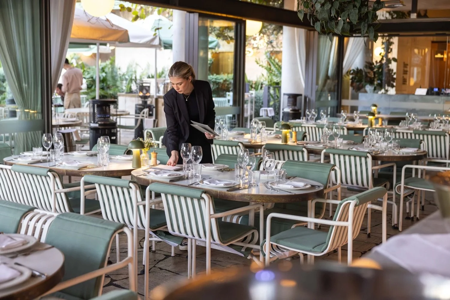 A woman setting a table in a stylish restaurant with green chairs and wine glasses, with outdoor patio visible through large glass windows and doors.