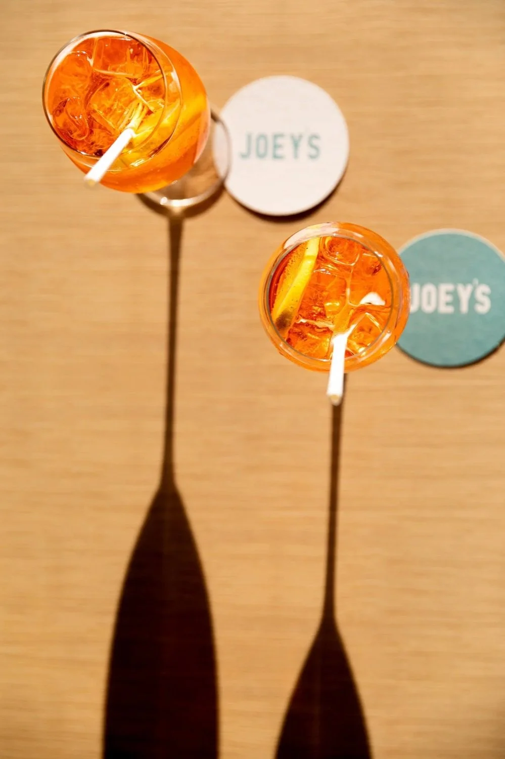 Two glasses of orange-colored drink with ice and lemon slices, placed on a wooden table with Joey's coasters.