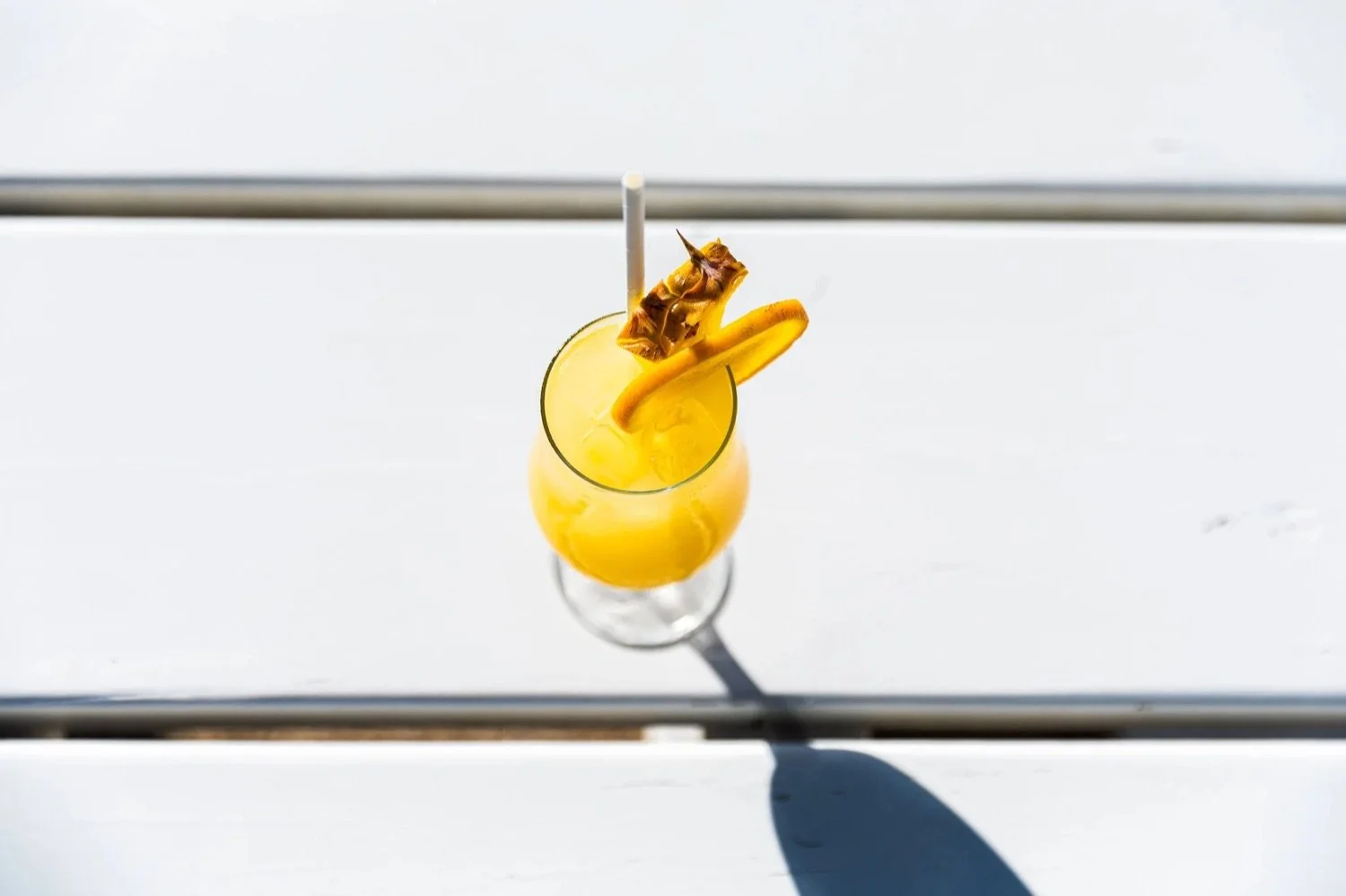 A tall glass of yellow tropical cocktail garnished with a pineapple slice, a small piece of pineapple, and a peel of orange, with a straw, on a white surface.