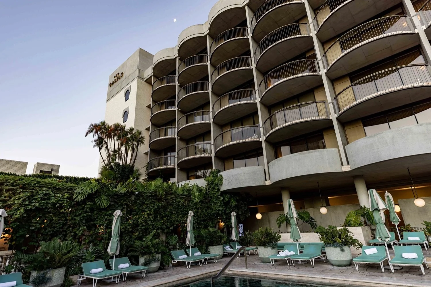 A beachfront hotel with multiple floors, balconies, and a pool area surrounded by green plants and lounge chairs, with umbrellas and lighting at sunset.