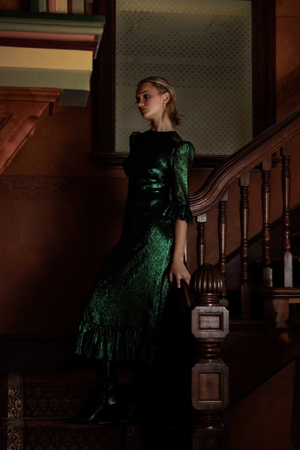 A woman with blonde hair standing on a wooden staircase, wearing a shiny green dress and black boots, inside a room with wooden walls and a bookshelf.