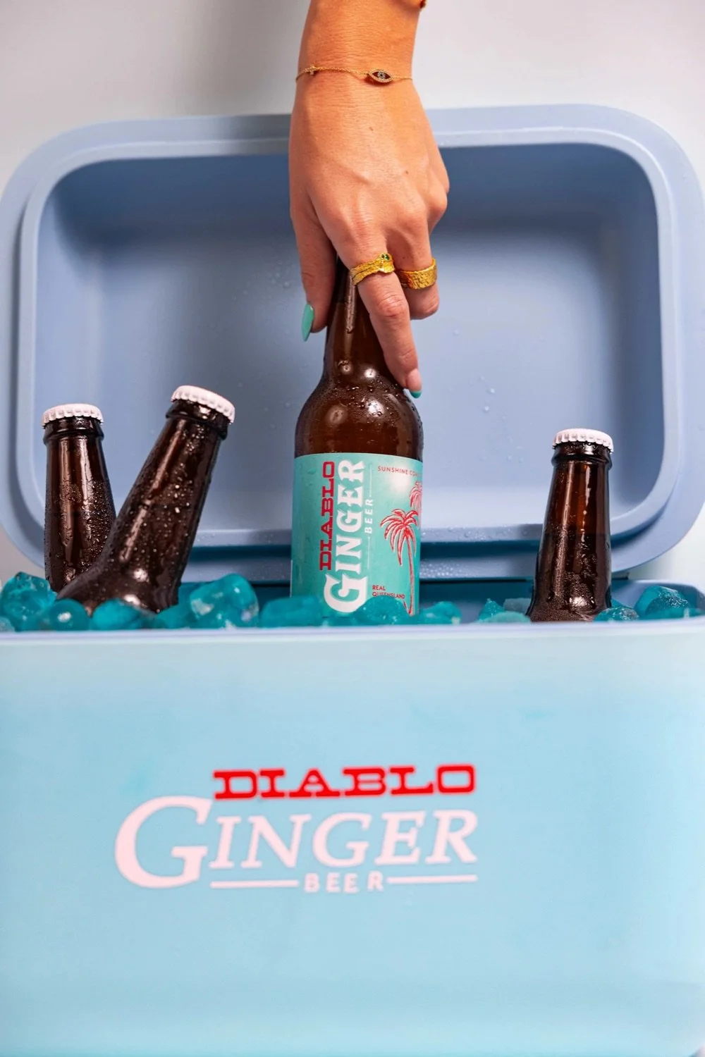 A hand with rings and a bracelet reaching into a freezer to grab a bottle of Diablo Ginger Beer, surrounded by other bottles in ice inside a light blue cooler.