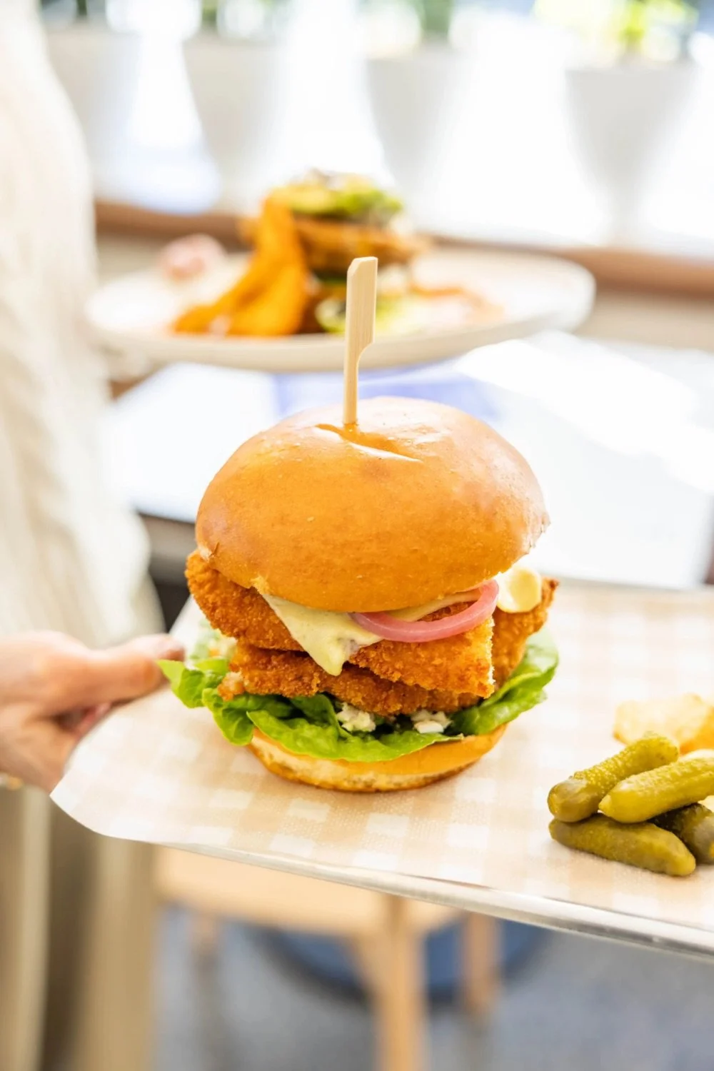 A person holding a sandwich with fried chicken fillet, lettuce, pickles, mayonnaise, and a bun, with a side of pickles on a white tray.