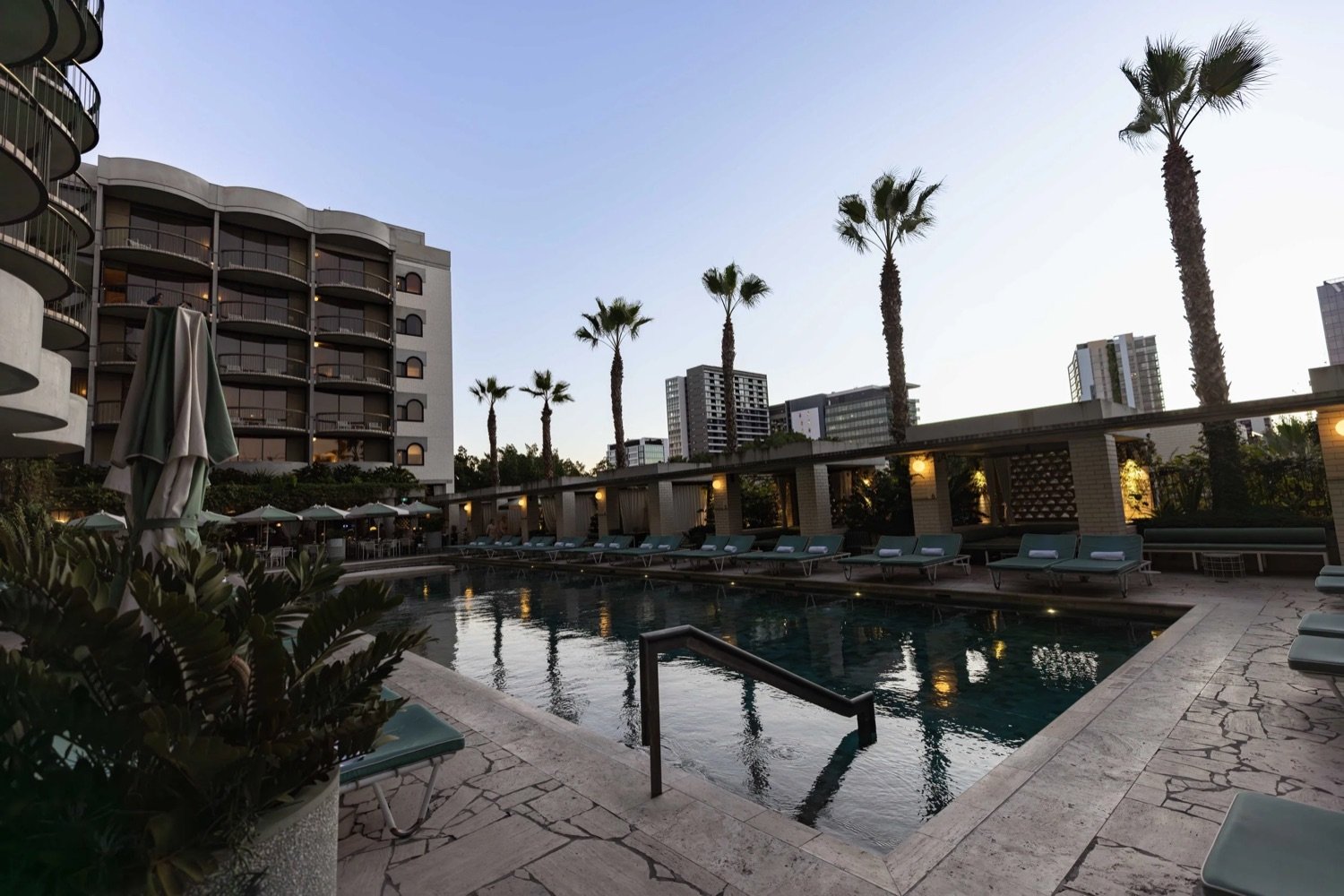 Poolside area with lounge chairs and umbrellas, tall palm trees, high-rise buildings in the background, nighttime lighting.