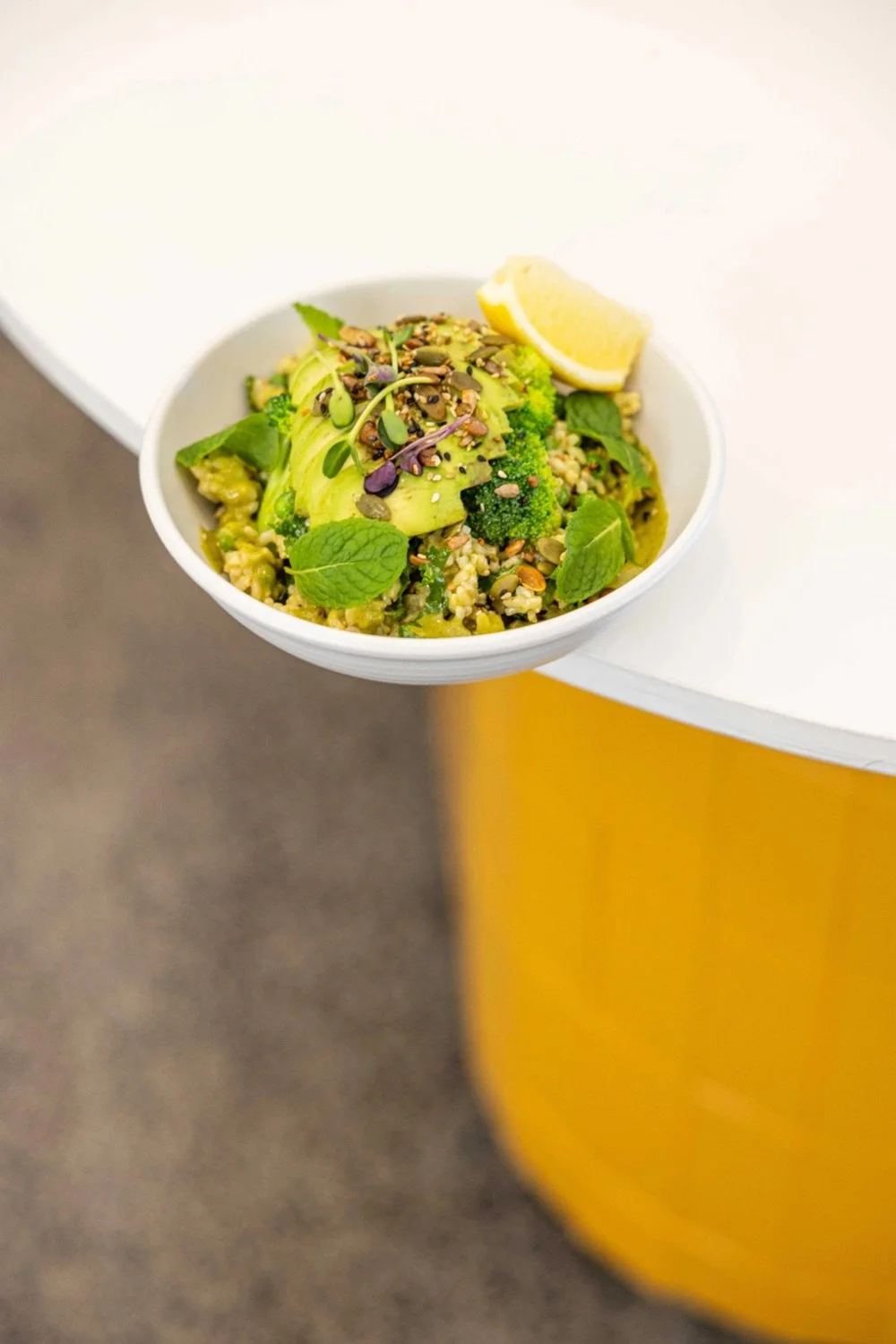 A bowl of grain salad topped with sliced avocado, microgreens, sunflower seeds, and lemon wedge on a white circular table.