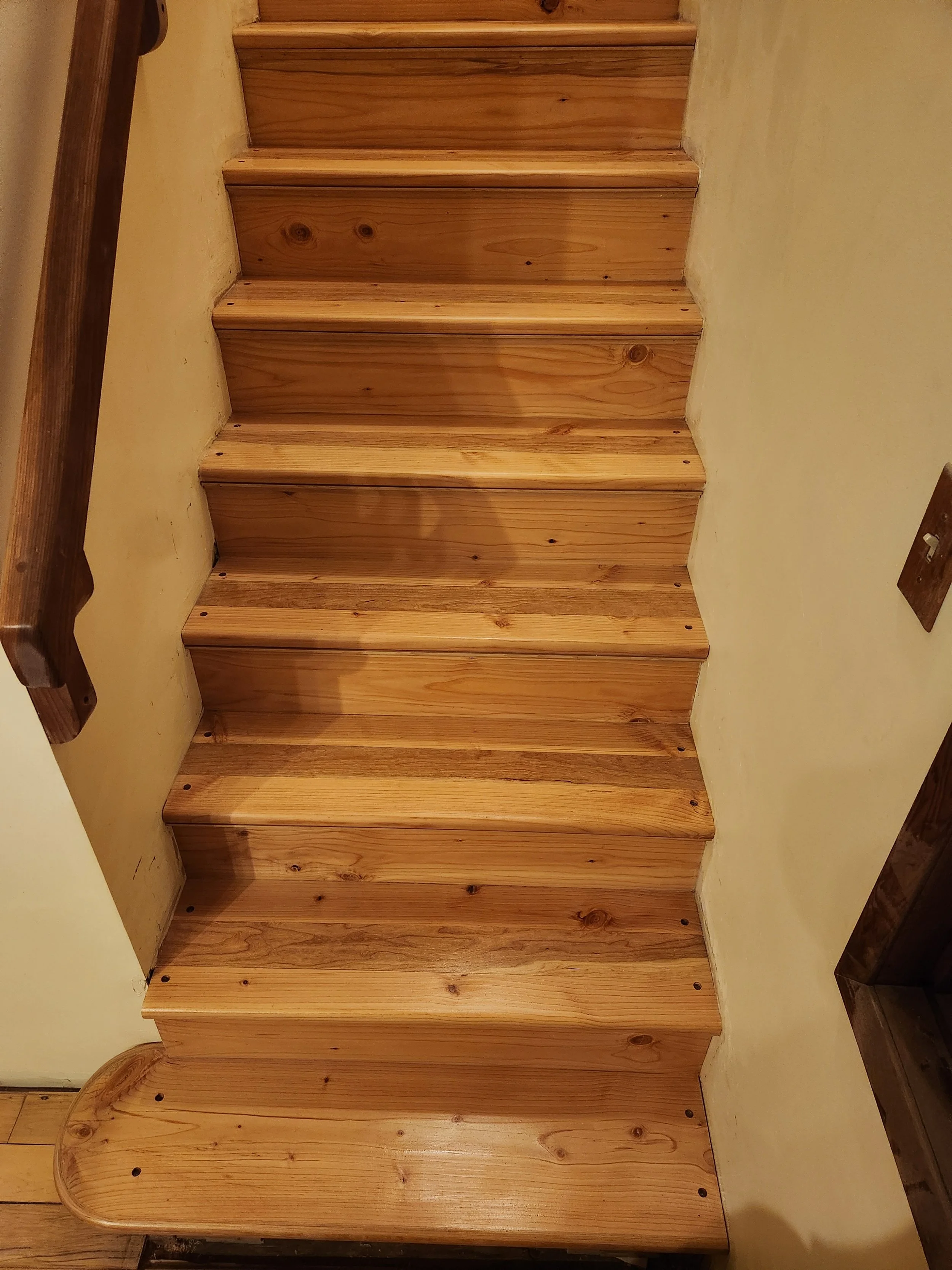 Wooden staircase ,viewed from below, with a handrail on the left side and a beige wall on the right.