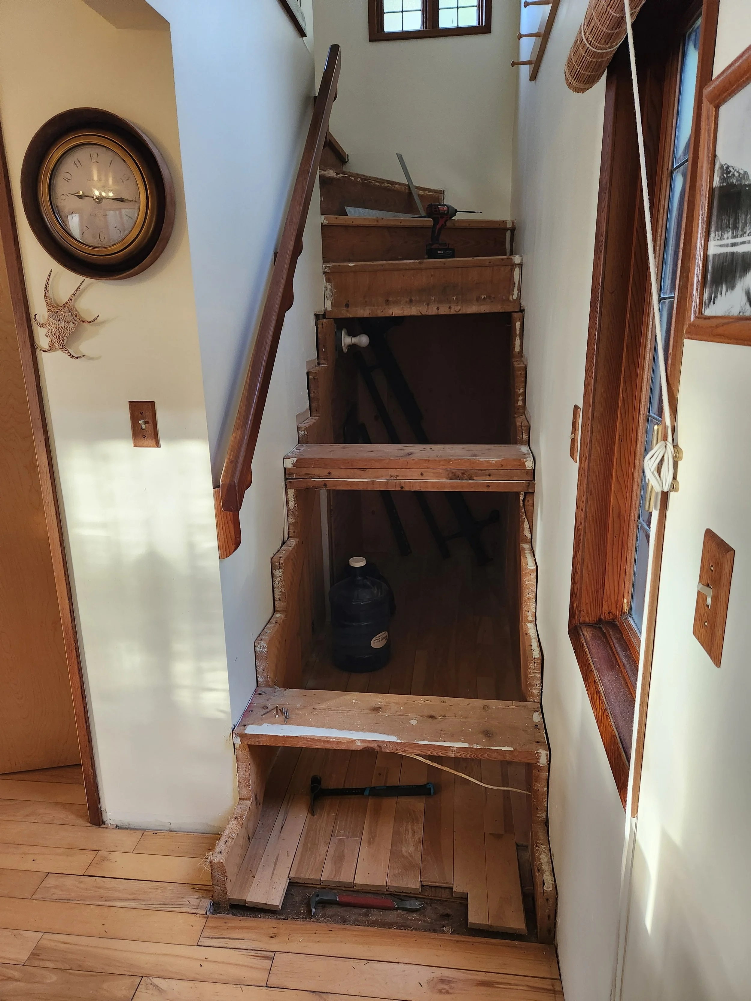 A staircase under renovation, with some steps removed, revealing the space underneath. The area contains tools like a hammer and a power drill, and a large water jug. The staircase and surrounding walls are partly finished, with visible wooden framing and steps in progress. A window on the right lets in natural light.