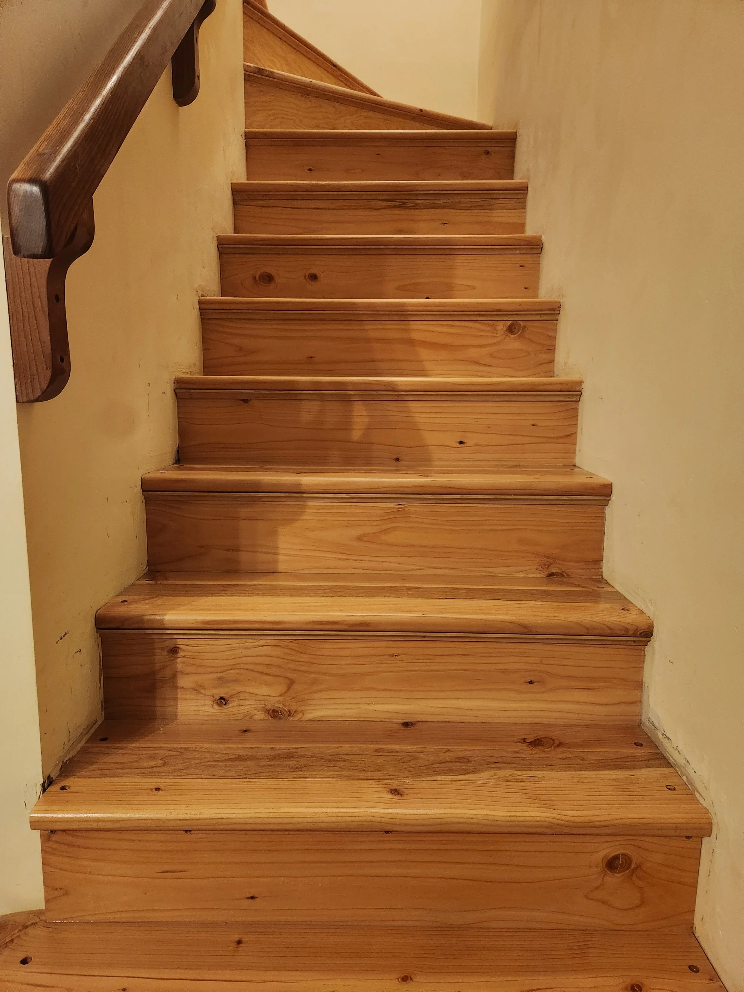 A wooden staircase with a handrail on the left side, leading upward against cream-colored walls.