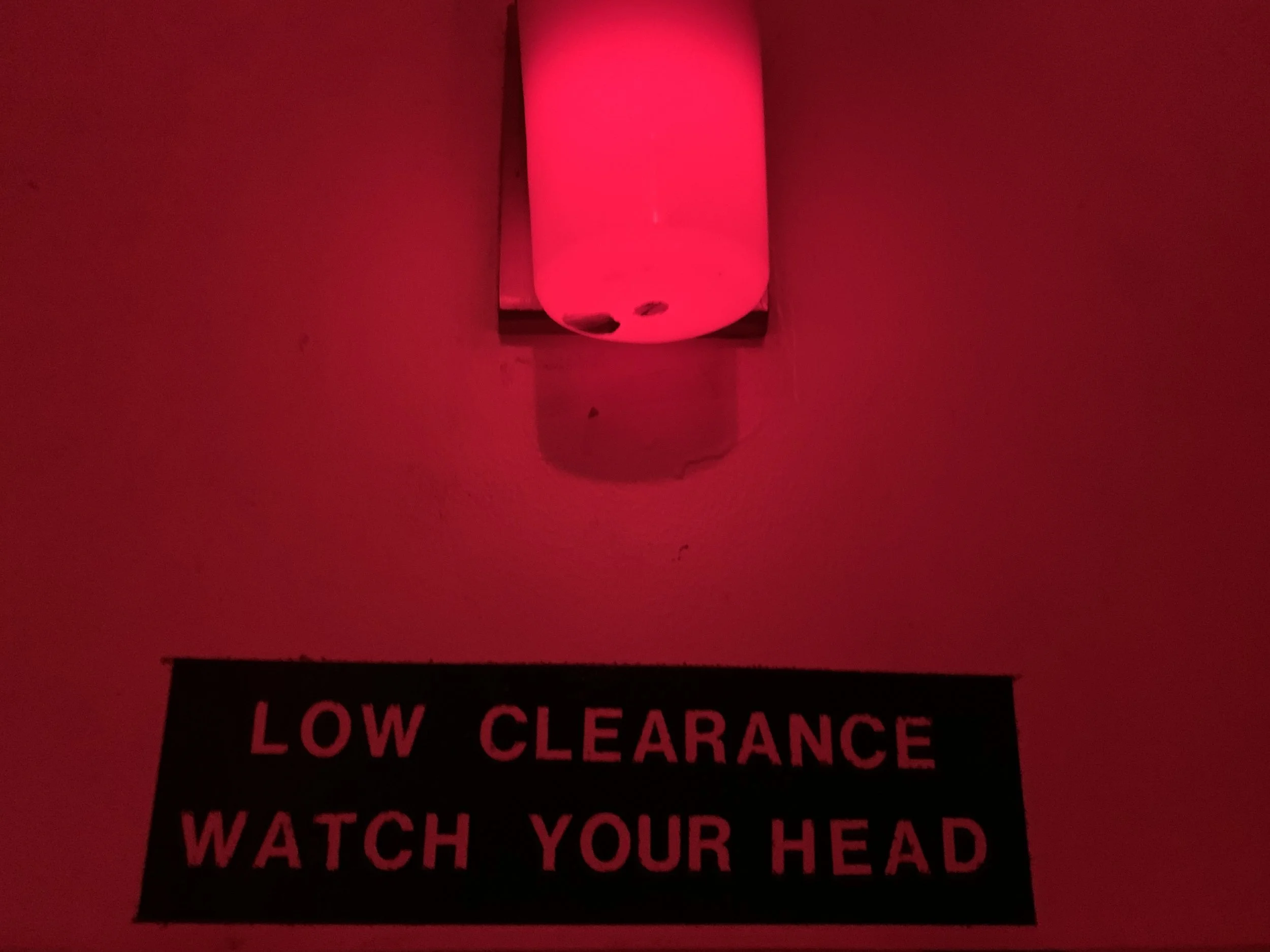 A red light fixture mounted on a wall above a warning sign that reads 'Low Clearance Watch Your Head'.