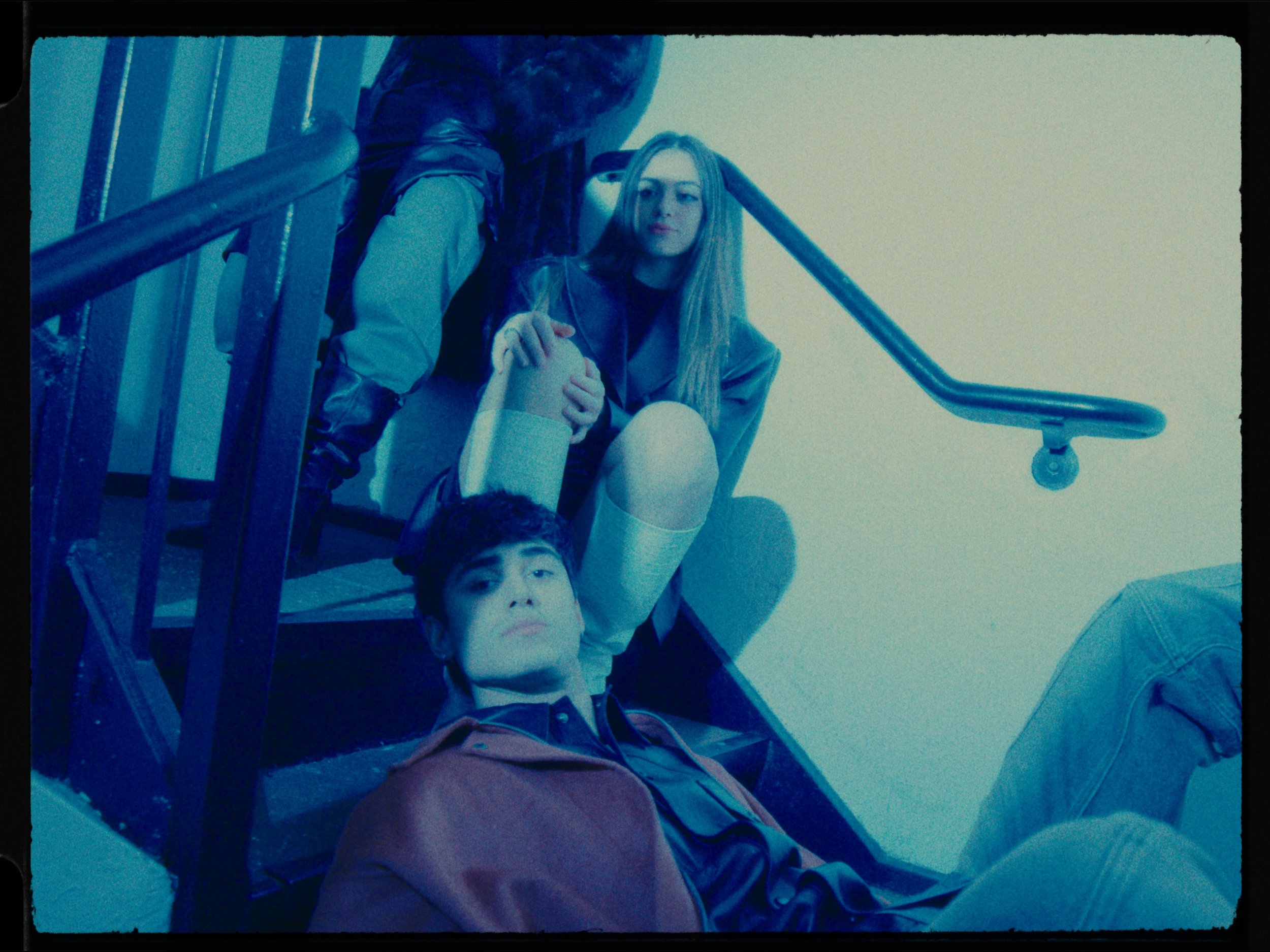 A young man with dark hair lying on the floor looking up, a young woman with long blonde hair sitting on stairs, and an adult woman with long hair and a patterned jacket leaning over the stairs.