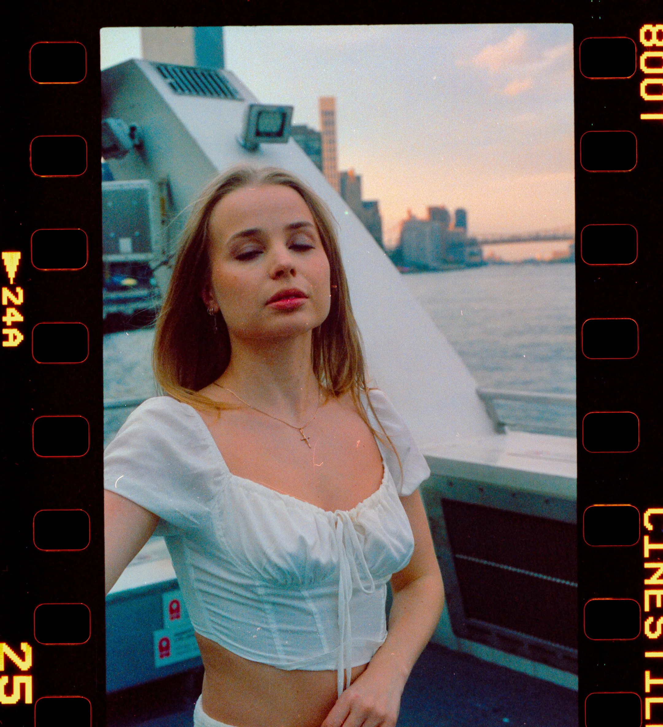 A woman with shoulder-length blonde hair wearing a white crop top poses on a boat with a city skyline and a river in the background during sunset.