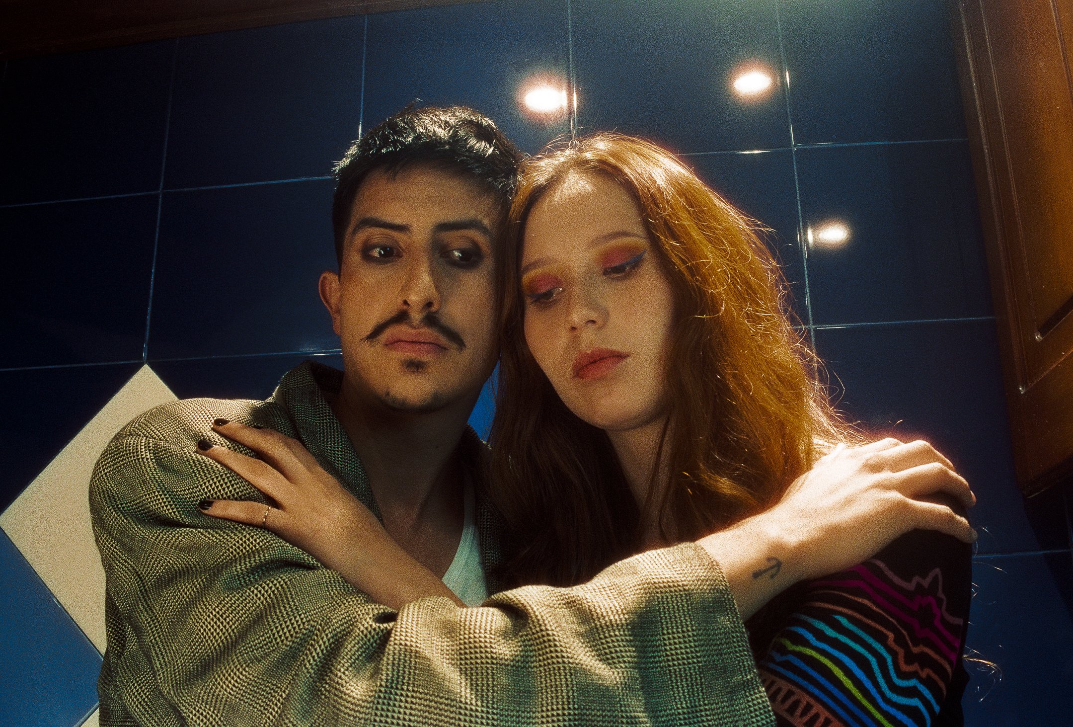 Two young adults, a man with short dark hair, a mustache and beard, and a woman with long red hair and colorful makeup, embracing each other in front of a blue tiled wall.