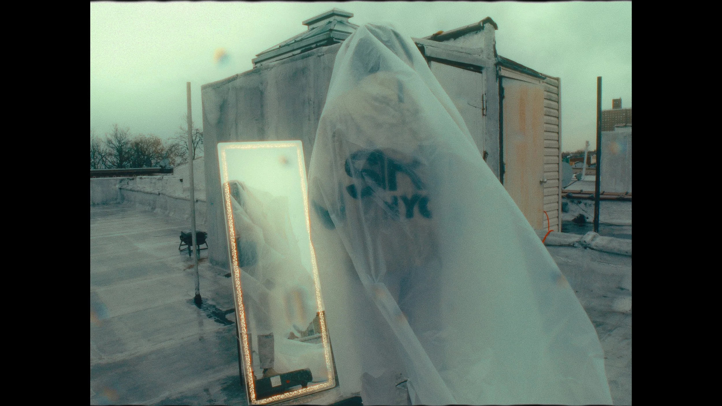 A person is seen taking a photo of themselves in a rectangular mirror with illuminated edges, on a rooftop. The reflection shows the person holding a phone, wearing a hoodie with the Nike logo. The scene has a cloudy sky and some rooftop structures.