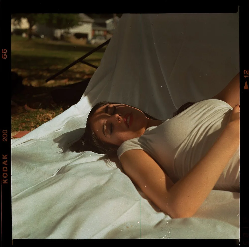 A woman is lying down on a white sheet outdoors, with her eyes closed and a slight smile, under a tent or canopy in a park or garden setting.