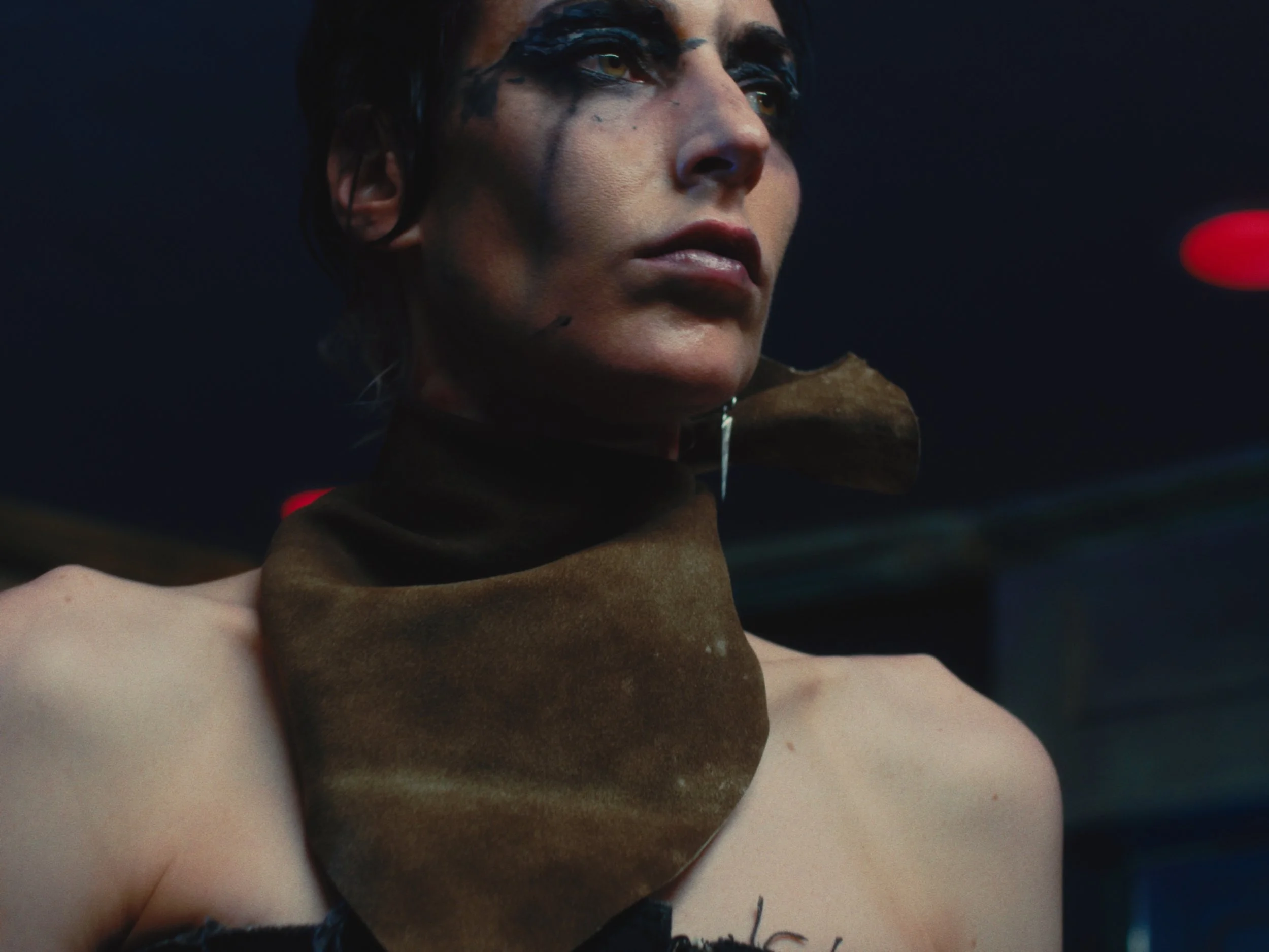 Close-up of a person with short dark hair, wearing dark makeup, a brown scarf around their neck, and no shirt, with a dark background and red lights in the distance.