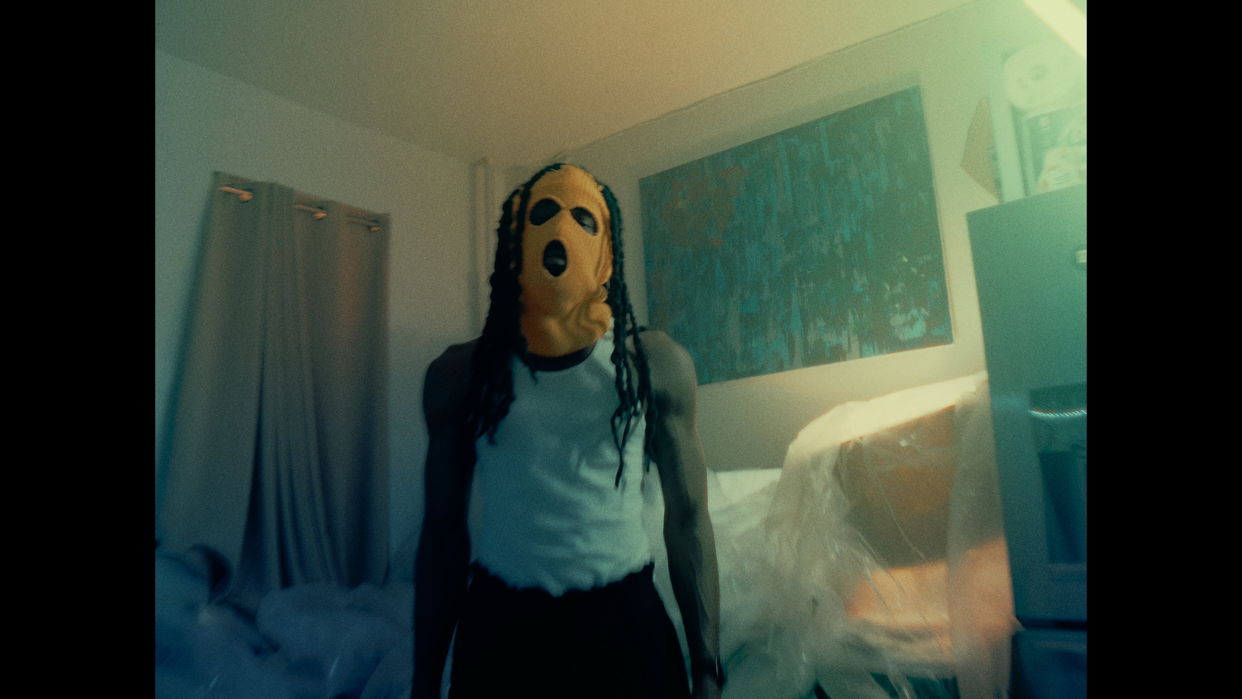 person wearing a yellow mask with black holes for eyes and mouth, with dreadlocks, standing in a dimly lit room with a bed, painting, and curtains.