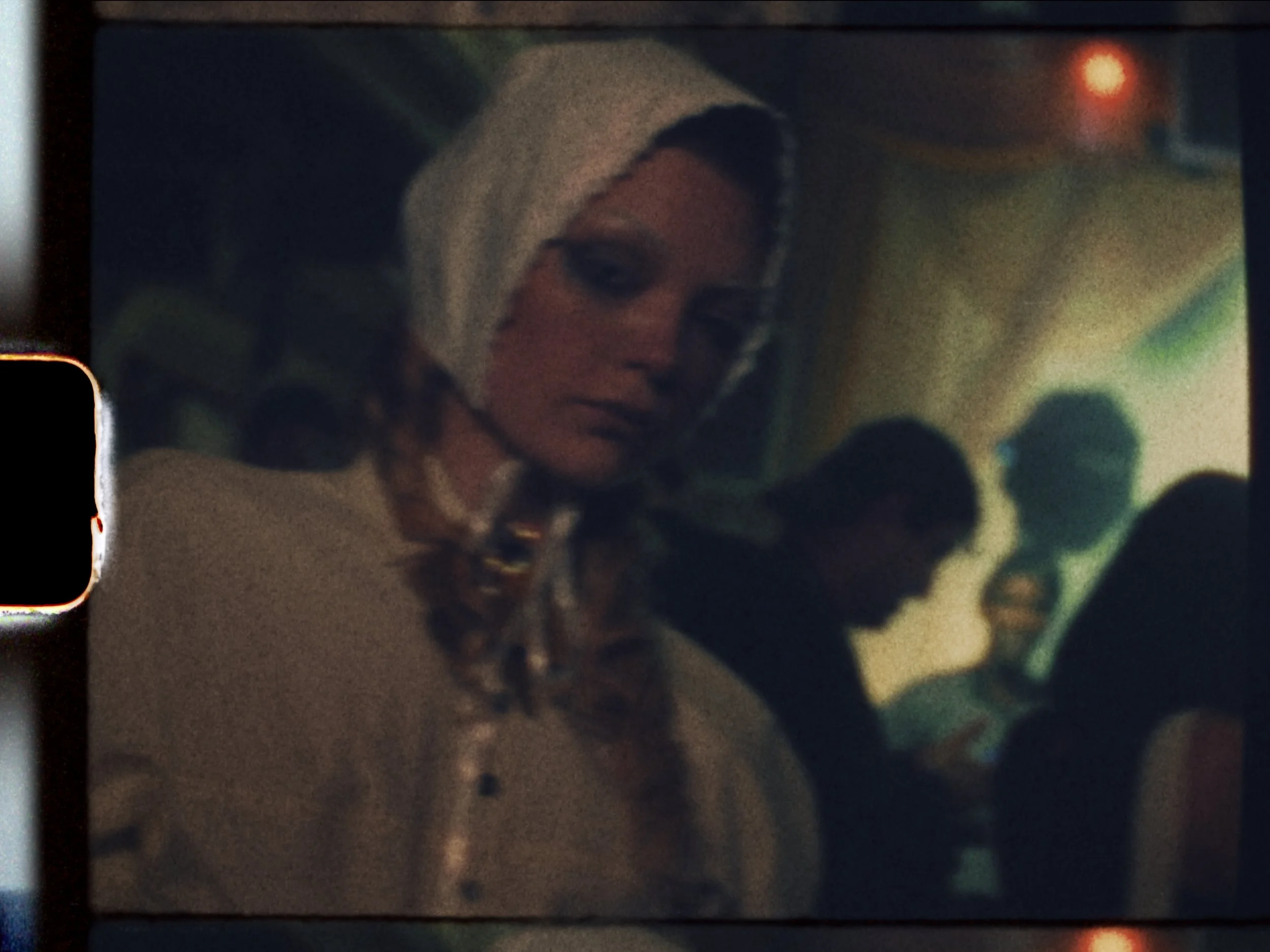 A woman with blonde hair and a beige hat looks over her shoulder in a dimly lit, crowded setting with other people in the background.