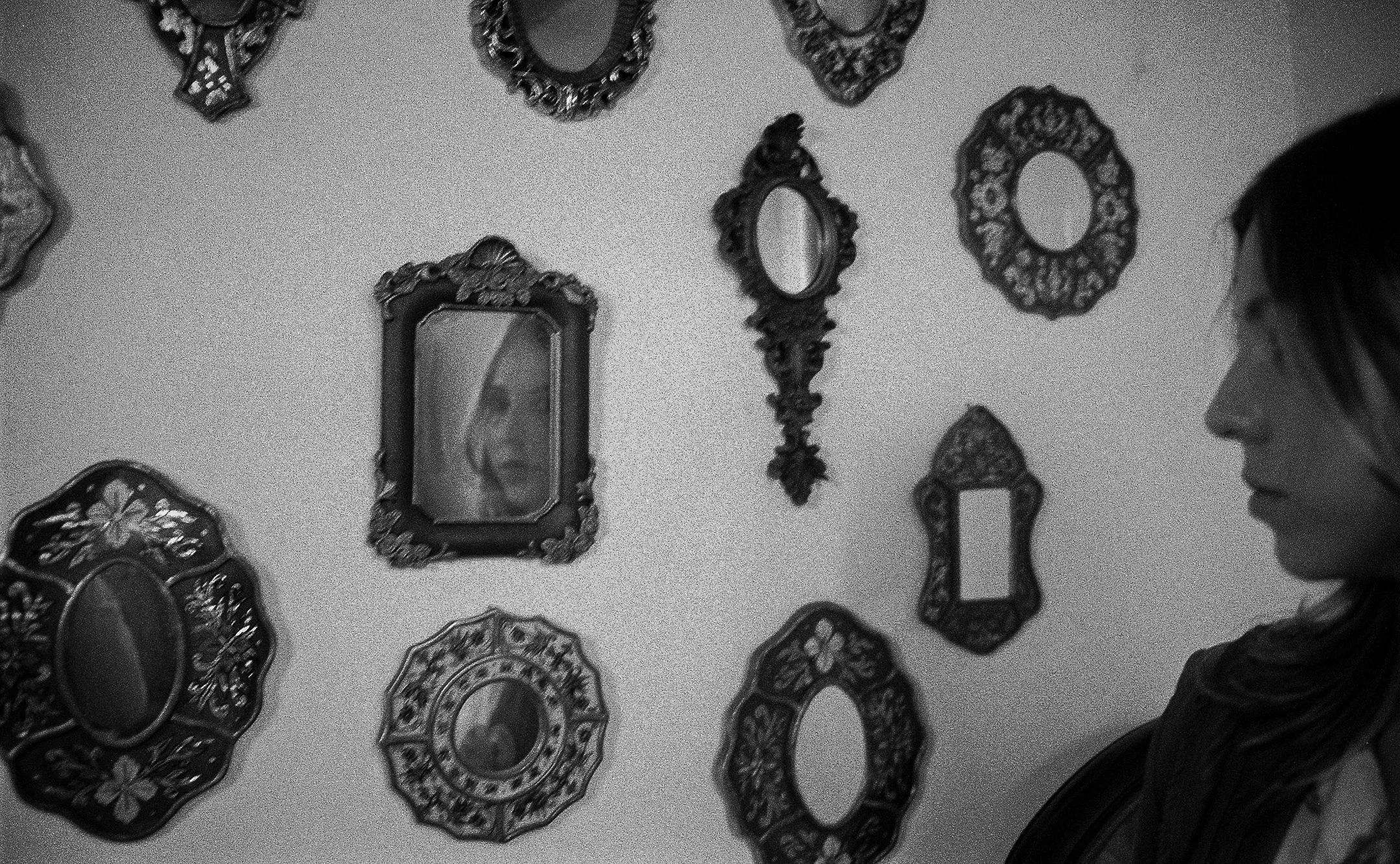 A woman with glasses on the right side of the frame looking at a wall decorated with multiple ornate picture frames and decorative mirrors.