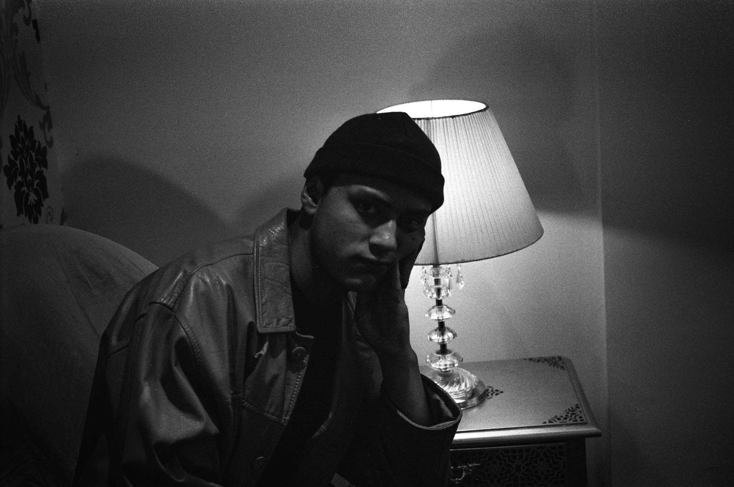A young man wearing a dark beanie and leather jacket sitting at a bedside table with a lamp, looking at the camera in a dimly lit room.