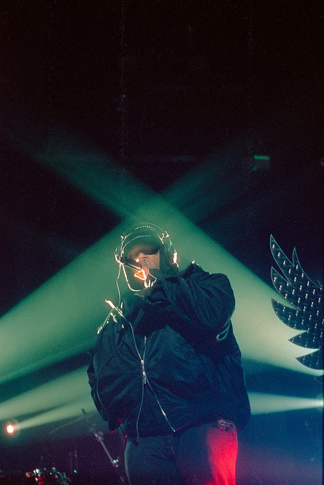 A performer wearing a motorcycle helmet and headphones, holding a microphone, illuminated by green stage lights with dark background.