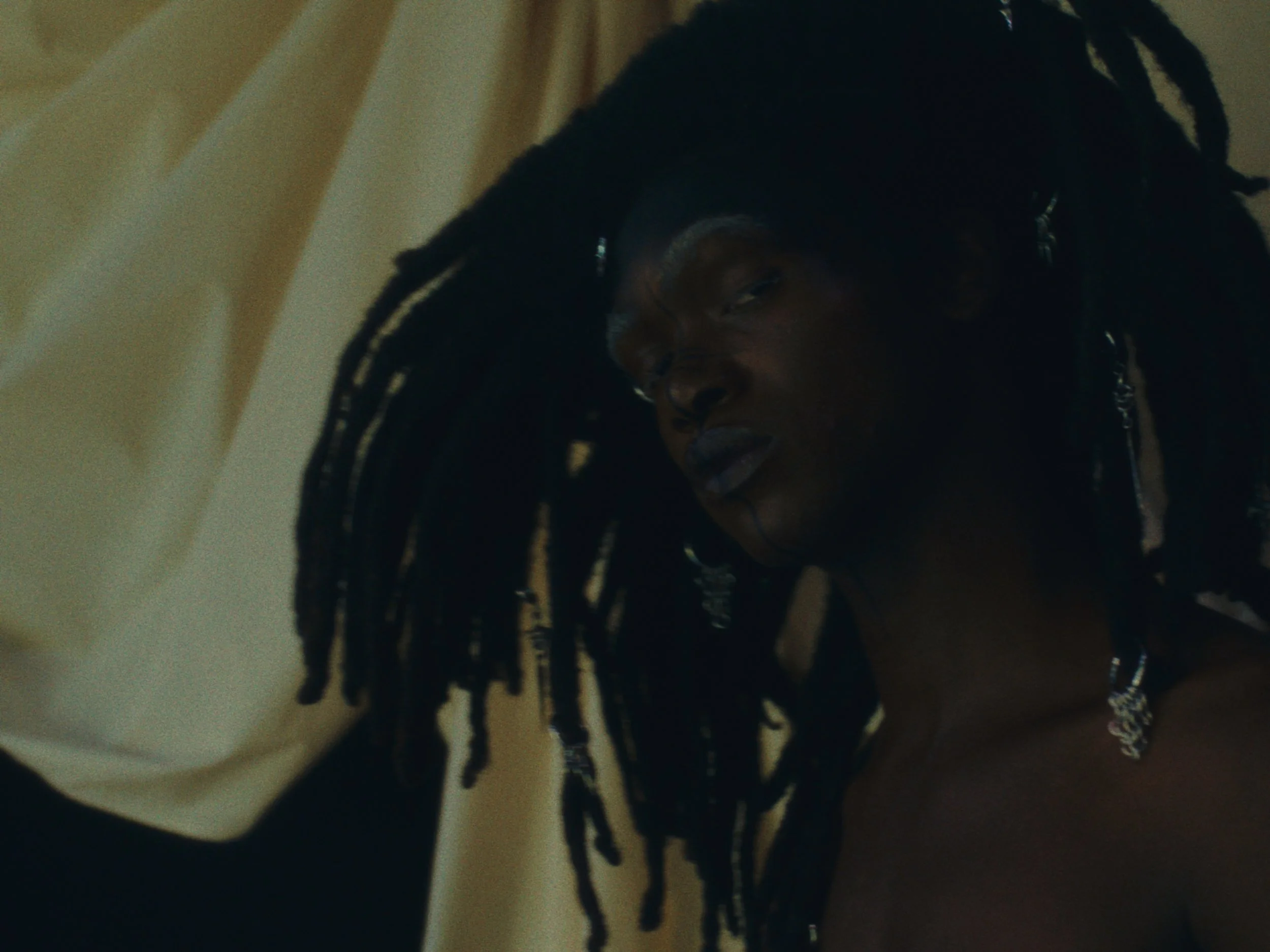 A person with long dreadlocks resting their head against a cream-colored pillow or surface, with a dark background.