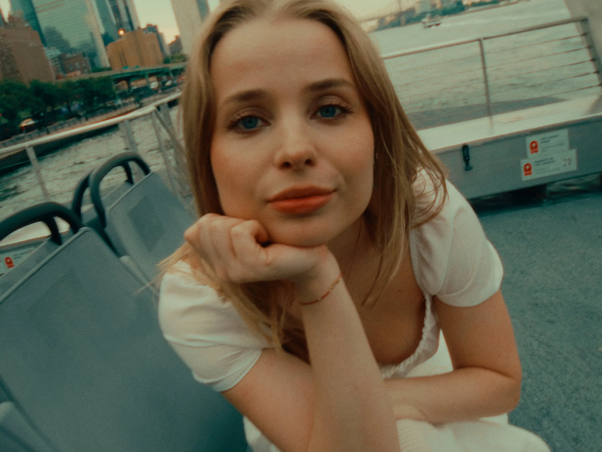 Close-up of a woman with blonde hair and blue eyes resting her chin on her hand on a boat with city skyline and water in the background.